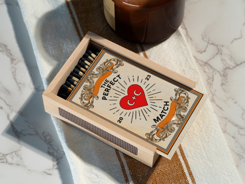 Puede incluir: Una caja de cerillas de madera con un dise&ntilde;o vintage que presenta un coraz&oacute;n con el texto "C+C" y las palabras "The Perfect Match 20 23". La caja de cerillas est&aacute; abierta y muestra una pila de cerillas.