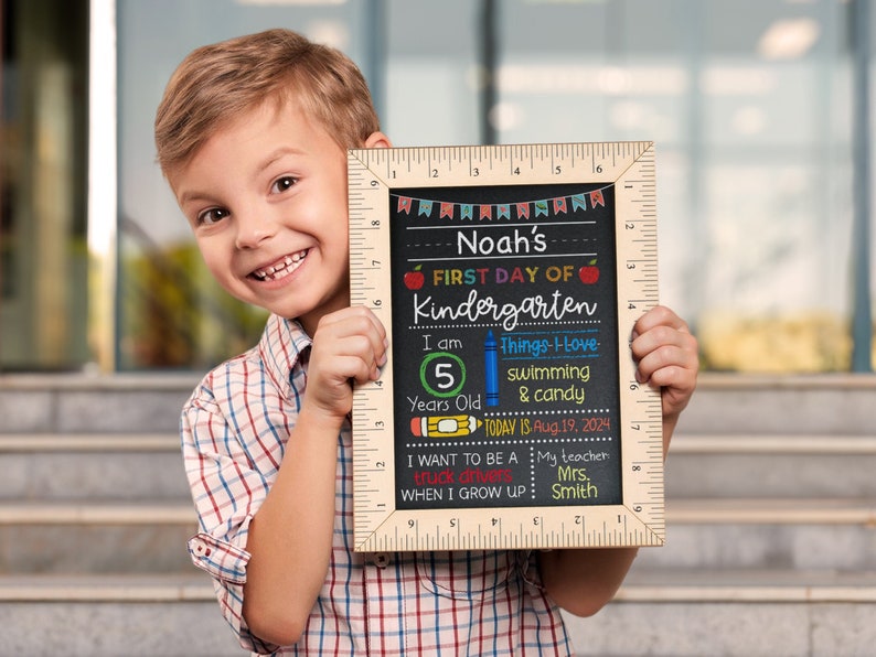 Puede incluir: Un letrero de pizarra con un marco de madera y una regla en el lateral. El letrero dice "Noah's First Day of Kindergarten". Tambi&eacute;n dice "I am 5 years old", "Things I love: swimming & candy", "Today is Aug. 19, 2024", "I want to be a truck driver when I grow up", y "My teacher Mrs. Smith".