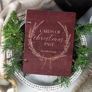 May include: A burgundy wooden scrapbook with a gold inscription that reads "Cards of Christmas Past" and "The Miller Family" surrounded by a floral wreath.