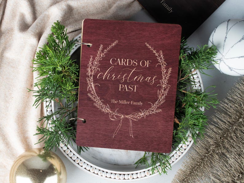 May include: A burgundy wooden scrapbook with a gold inscription that reads "Cards of Christmas Past" and "The Miller Family" surrounded by a floral wreath.