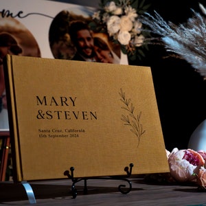 Puede incluir: Un libro de visitas de boda dorado con los nombres "MARY & STEVEN" impresos en negro. El libro tambi&eacute;n incluye la fecha y el lugar de la boda, y una ramita decorativa. El libro se muestra en un soporte de metal negro.
