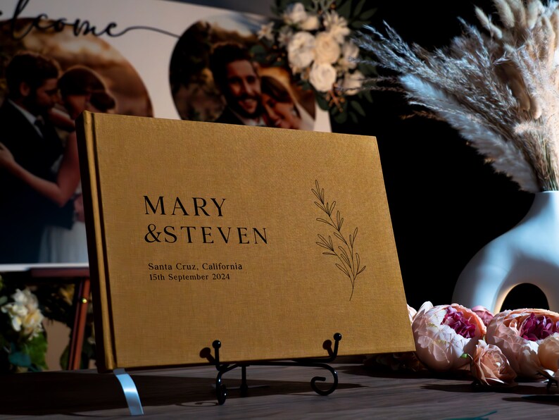 Puede incluir: Un libro de visitas de boda dorado con los nombres "MARY & STEVEN" impresos en negro. El libro tambi&eacute;n incluye la fecha y el lugar de la boda, y una ramita decorativa. El libro se muestra en un soporte de metal negro.