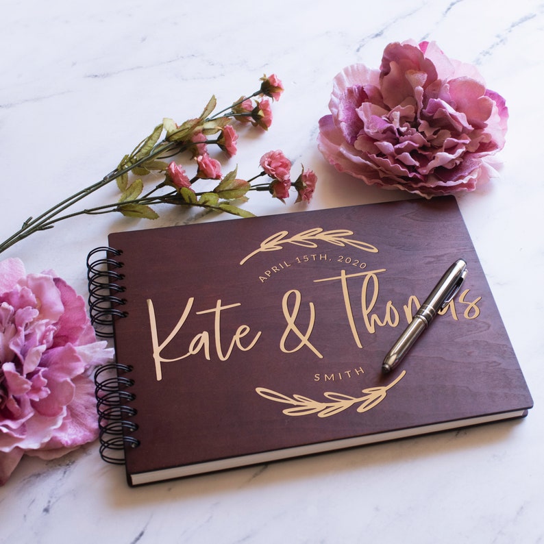 May include: A dark brown wooden guest book with a gold inscription that reads "Kate & Thomas Smith, April 15th, 2020". The book is open and a silver pen is lying on top of it.