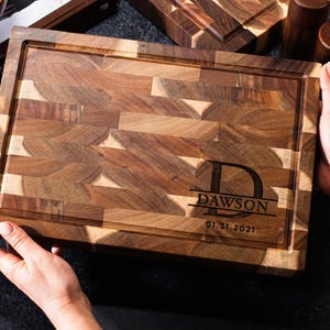 May include: A rectangular wooden chopping board with a juice groove and a personalized engraving. The board has a mosaic pattern of various wood tones and the name "Dawson" with a date. The board is held by two hands.