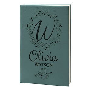 May include: Teal leather-bound journal with a floral wreath design and the initial "W" in the center. The name "Olivia Watson" and the year "2020" are engraved below the wreath.