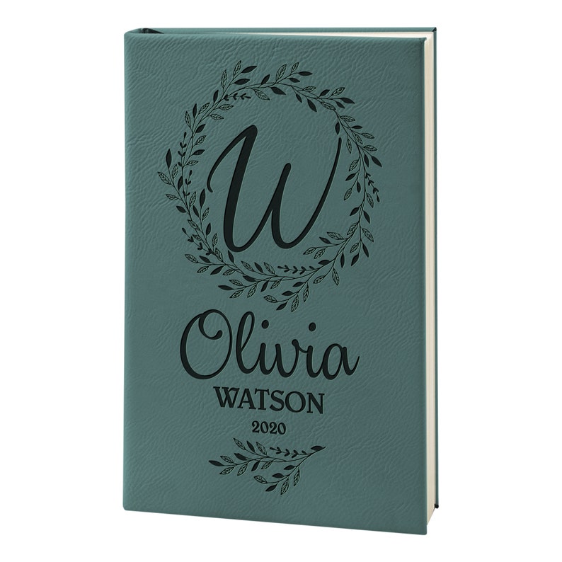May include: Teal leather-bound journal with a floral wreath design and the initial "W" in the center. The name "Olivia Watson" and the year "2020" are engraved below the wreath.