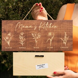 Puede incluir: Un letrero de madera con el texto "Mama's Kitchen" y cinco dise&ntilde;os florales con nombres debajo de cada dise&ntilde;o: Elijah, Sophia, Noah, Clair y Lindsay. El letrero est&aacute; hecho a mano en los Estados Unidos.
