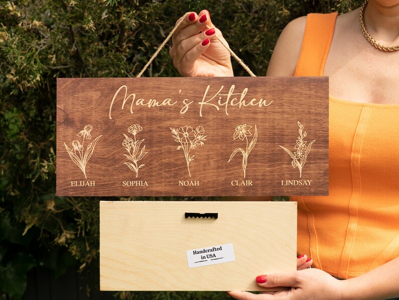 Puede incluir: Un letrero de madera con el texto "Mama's Kitchen" y cinco dise&ntilde;os florales con nombres debajo de cada dise&ntilde;o: Elijah, Sophia, Noah, Clair y Lindsay. El letrero est&aacute; hecho a mano en los Estados Unidos.