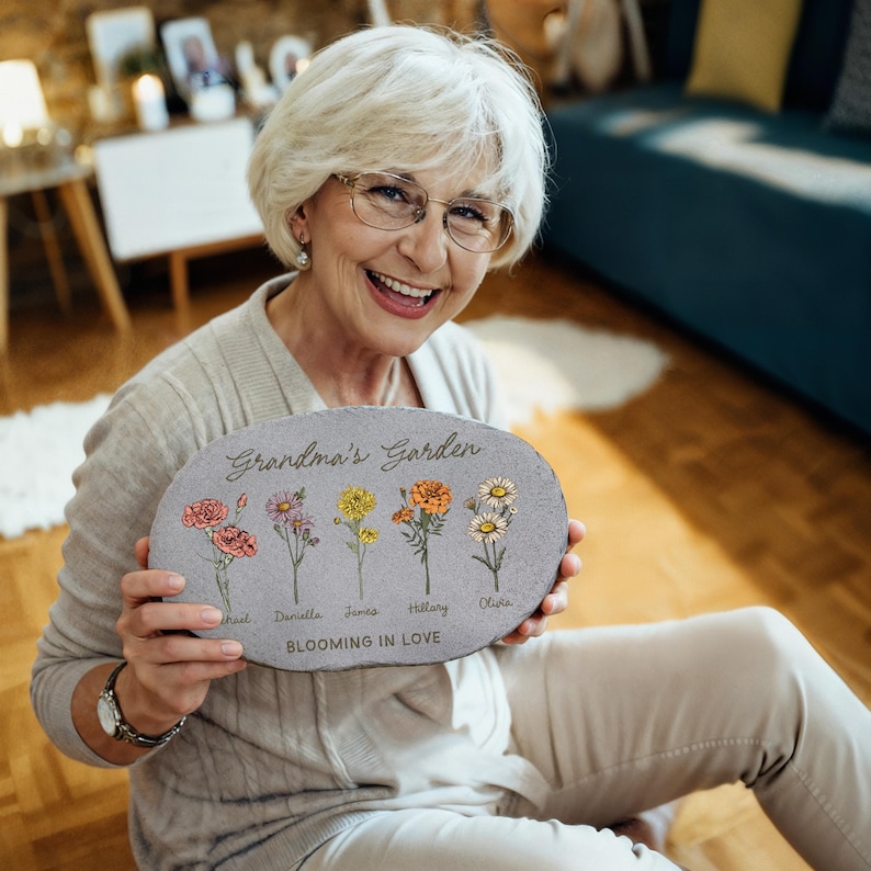 May include: An oval gray stone garden plaque with the text "Grandma's Garden" and the words "Blooming in Love." The plaque features illustrations of five different flowers with names below each flower. The woman in the image is smiling.