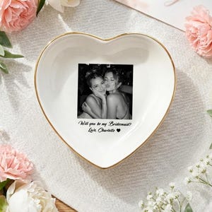 May include: A white heart-shaped ceramic trinket dish with a gold-colored rim. The center features a black and white photograph of two people and the printed text: Will you be my Bridesmaid? Love, Charlotte with a small black heart symbol.