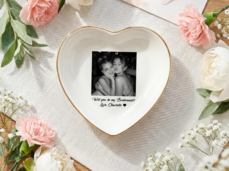 May include: A white heart-shaped ceramic trinket dish with a gold-colored rim. The center features a black and white photograph of two people and the printed text: Will you be my Bridesmaid? Love, Charlotte with a small black heart symbol.
