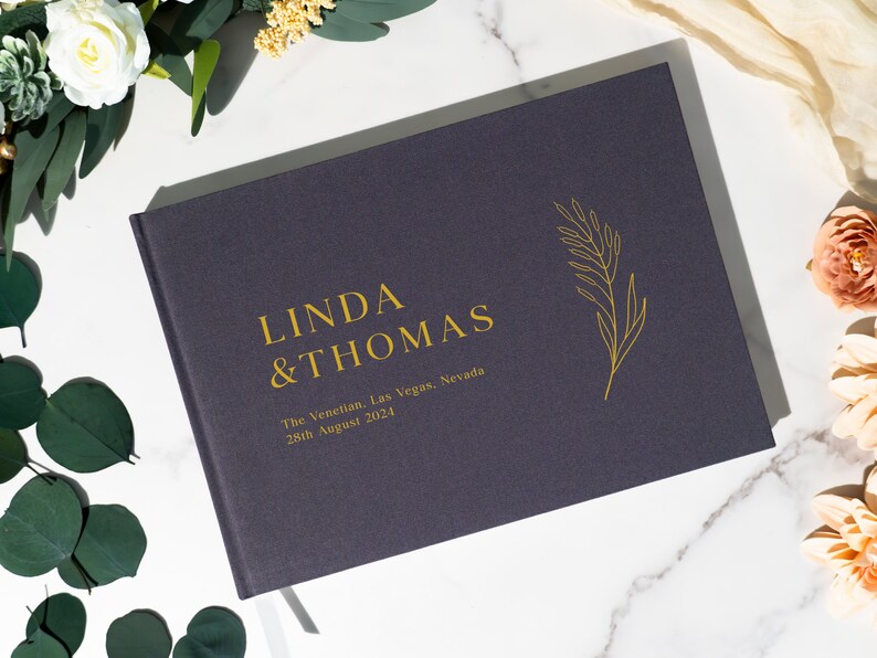 Puede incluir: Un libro de tapa dura azul marino con letras doradas que dice "LINDA & THOMAS". Debajo de los nombres est&aacute; "The Venetian, Las Vegas, Nevada 28th August 2021". Un dise&ntilde;o de hoja dorada est&aacute; a la derecha. El libro est&aacute; rodeado de flores.