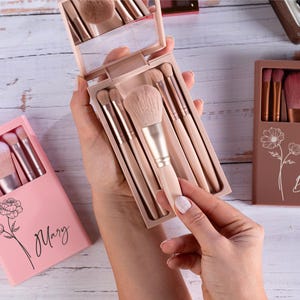 May include: A collection of makeup brush sets in pink and brown cases. The sets include various brushes with light-colored handles and soft bristles. One case is open, revealing a mirror and brushes. The cases are decorated with floral designs and names.