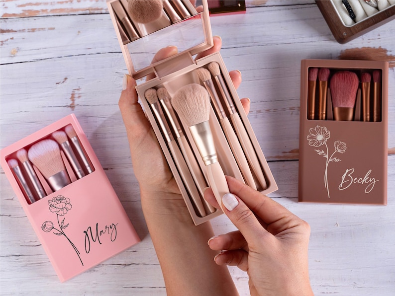 May include: A collection of makeup brush sets in pink and brown cases. The sets include various brushes with light-colored handles and soft bristles. One case is open, revealing a mirror and brushes. The cases are decorated with floral designs and names.