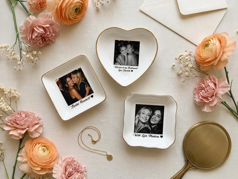 May include: Three white ceramic trinket dishes with gold-toned rims, featuring custom black and white photographs and printed text. One heart-shaped dish reads Will you be my Bridesmaid? Love Charlotte, one square dish reads Besties Forever!!, and one reads With Love, Madison.