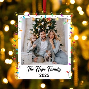 Pode incluir: Um enfeite de Natal em acr&iacute;lico transparente com uma foto de fam&iacute;lia no interior, emoldurado por uma s&eacute;rie de luzes coloridas. A foto mostra uma fam&iacute;lia de tr&ecirc;s pessoas e um c&atilde;o em frente a uma &aacute;rvore de Natal. O texto "The Hope Family 2025" est&aacute; na parte inferior.