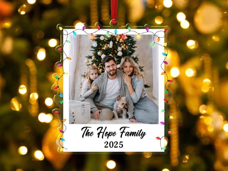 Pode incluir: Um enfeite de Natal em acr&iacute;lico transparente com uma foto de fam&iacute;lia no interior, emoldurado por uma s&eacute;rie de luzes coloridas. A foto mostra uma fam&iacute;lia de tr&ecirc;s pessoas e um c&atilde;o em frente a uma &aacute;rvore de Natal. O texto "The Hope Family 2025" est&aacute; na parte inferior.