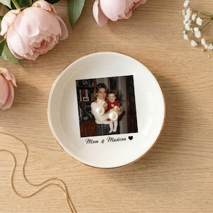 May include: A round white ceramic trinket dish with a gold-colored rim. The center features a printed photograph of an adult holding a child, with the text "Mom & Madison" and a small black heart symbol printed below the image.