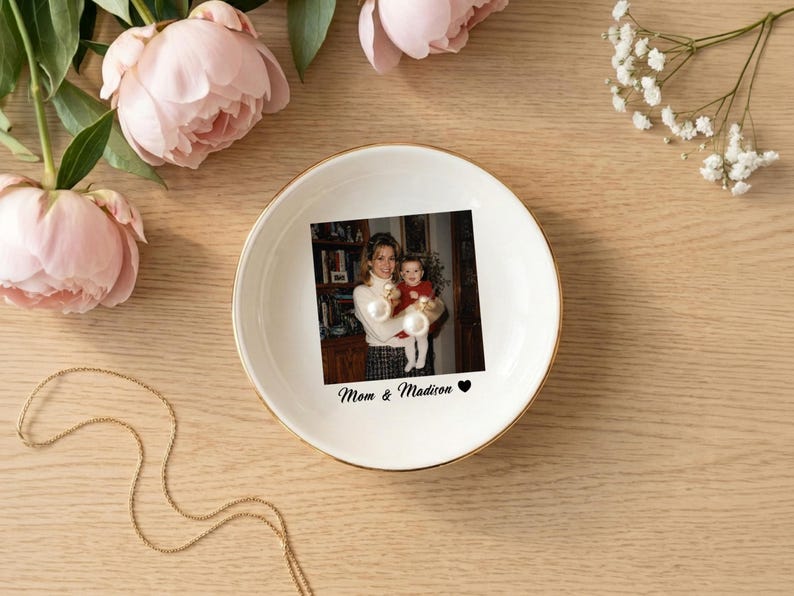May include: A round white ceramic trinket dish with a gold-colored rim. The center features a printed photograph of an adult holding a child, with the text "Mom & Madison" and a small black heart symbol printed below the image.