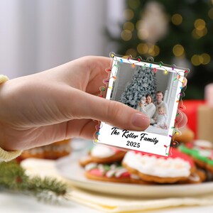 Pode incluir: Um enfeite de Natal em acr&iacute;lico transparente em forma de foto polaroid, segurado em uma m&atilde;o. A foto mostra uma fam&iacute;lia em frente a uma &aacute;rvore de Natal. O enfeite &eacute; decorado com luzes coloridas e o texto "The Keller Family 2025".
