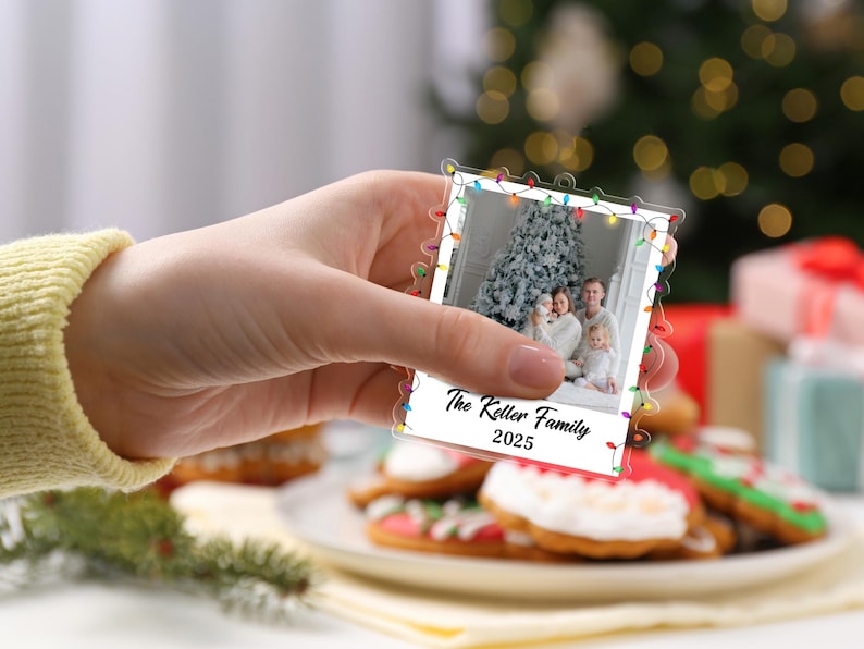Pode incluir: Um enfeite de Natal em acr&iacute;lico transparente em forma de foto polaroid, segurado em uma m&atilde;o. A foto mostra uma fam&iacute;lia em frente a uma &aacute;rvore de Natal. O enfeite &eacute; decorado com luzes coloridas e o texto "The Keller Family 2025".