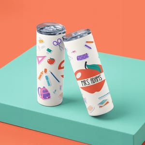 May include: Two white insulated tumblers with school-themed stickers. One tumbler features an apple graphic with the text "MRS. ADAMS". The other tumbler has school supplies stickers. Both tumblers have silver lids.