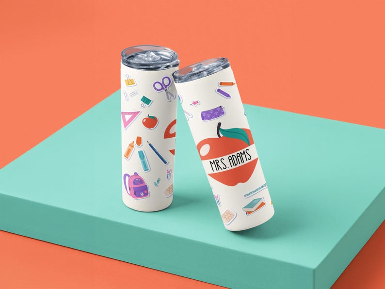 May include: Two white insulated tumblers with school-themed stickers. One tumbler features an apple graphic with the text "MRS. ADAMS". The other tumbler has school supplies stickers. Both tumblers have silver lids.