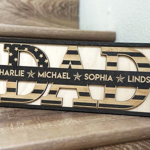 May include: Wooden sign with the word "DAD" in large letters, each letter is decorated with an American flag pattern. A smaller banner below the letters reads "CHARLIE MICHAEL SOPHIA LINDSAY".