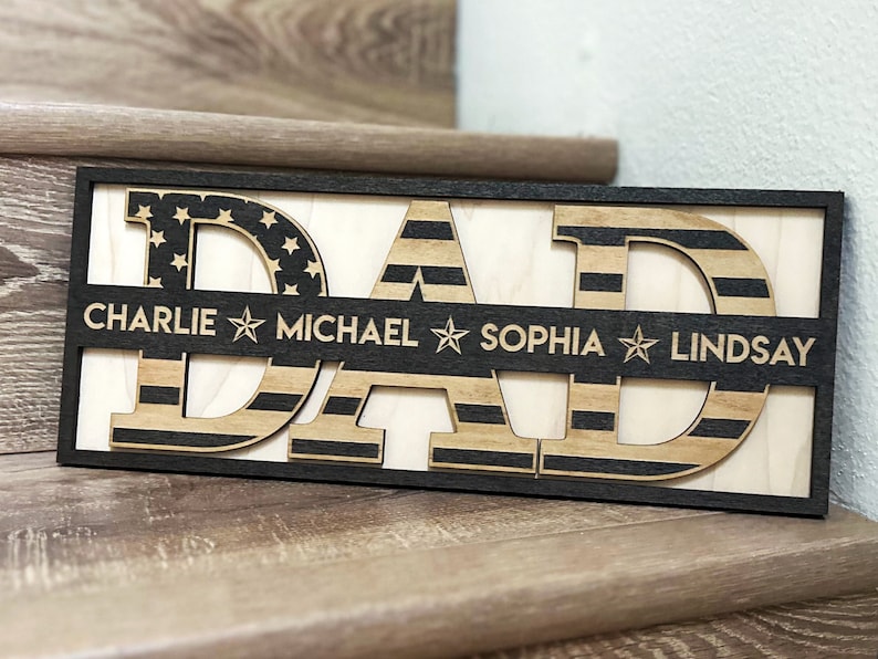 May include: Wooden sign with the word "DAD" in large letters, each letter is decorated with an American flag pattern. A smaller banner below the letters reads "CHARLIE MICHAEL SOPHIA LINDSAY".