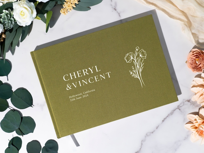 Puede incluir: Libro de visitas de boda verde oliva con los nombres "Cheryl & Vincent" en letras blancas. El libro tambi&eacute;n incluye el lugar y la fecha de la boda. Un dise&ntilde;o floral blanco est&aacute; en el lado derecho.