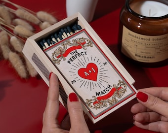 May include: A rectangular wooden matchbox featuring a label with the text THE PERFECT MATCH, a central red heart with the letters A+J, and the numbers 20 and 26. The box contains matches with dark blue heads and has a textured striking strip on the side.