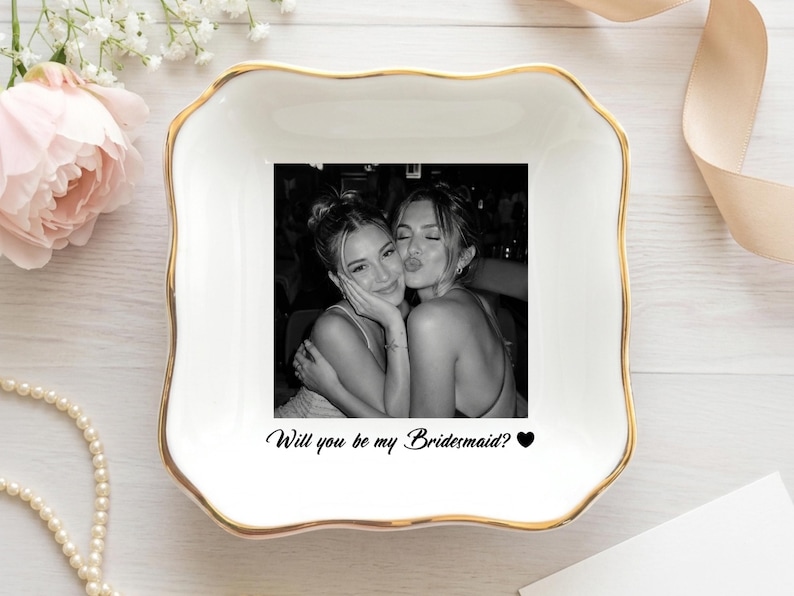 May include: A square white ceramic trinket dish with scalloped edges and a gold-painted rim. The center features a black and white photograph of two people embracing, with the black text "Will you be my Bridesmaid?" and a small heart icon below.