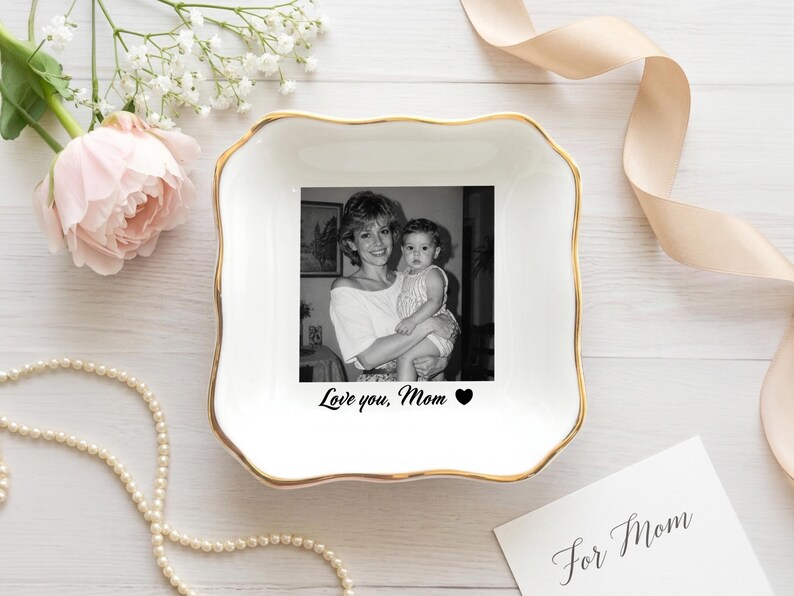 May include: A white ceramic trinket dish with a gold rim, featuring a black and white photo of a mother and child. The text "Love you, Mom" is below the photo. A pearl necklace and flowers are in the background.
