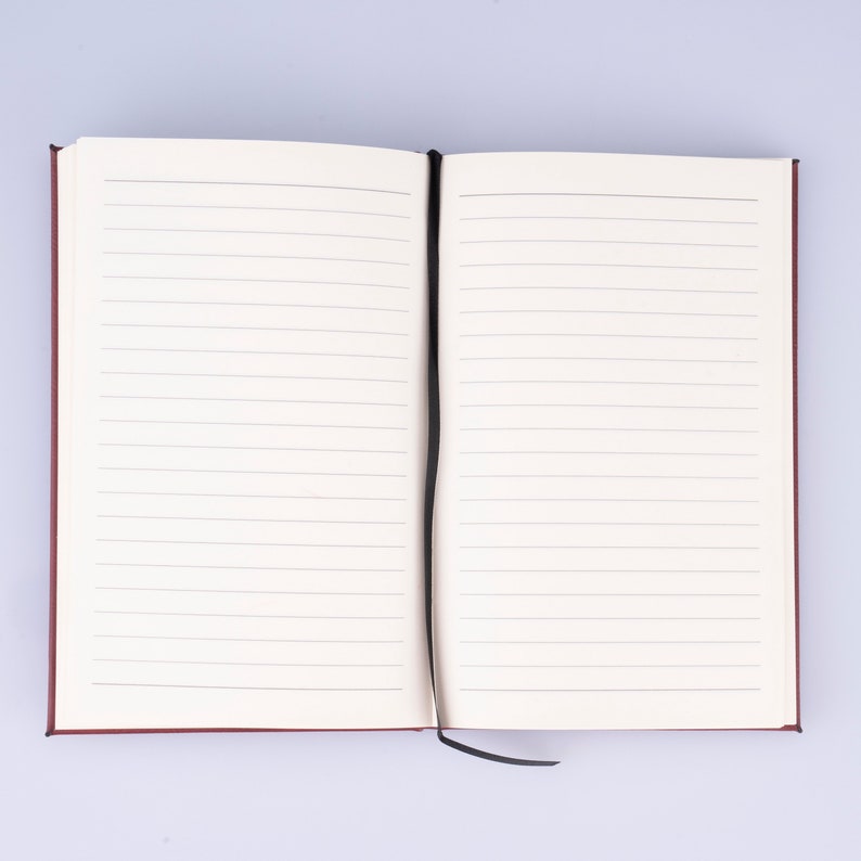 May include: A burgundy notebook with a black ribbon bookmark. The notebook is open to a blank page with lined paper.
