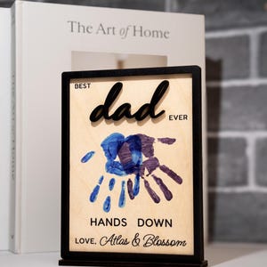 May include: A wooden plaque with a black frame and stand. The plaque reads "Best Dad Ever" with a handprint design in blue and purple. The text "Hands Down" and "Love, Atlas & Blossom" are also included.