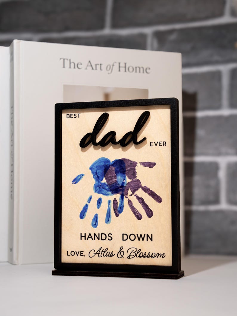 May include: A wooden plaque with a black frame and stand. The plaque reads "Best Dad Ever" with a handprint design in blue and purple. The text "Hands Down" and "Love, Atlas & Blossom" are also included.