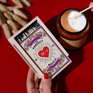Puede incluir: Una caja de madera de cerillas con un coraz&oacute;n rojo y el texto "The Perfect Match" y "M+J" en la parte delantera. La caja est&aacute; abierta y las cerillas son visibles. Una cerilla encendida se est&aacute; utilizando para encender una vela.