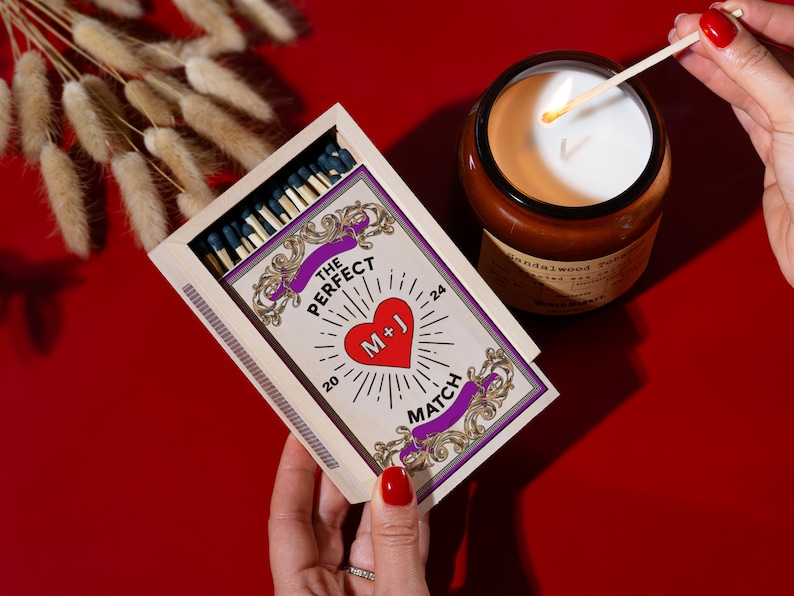 Puede incluir: Una caja de madera de cerillas con un coraz&oacute;n rojo y el texto "The Perfect Match" y "M+J" en la parte delantera. La caja est&aacute; abierta y las cerillas son visibles. Una cerilla encendida se est&aacute; utilizando para encender una vela.