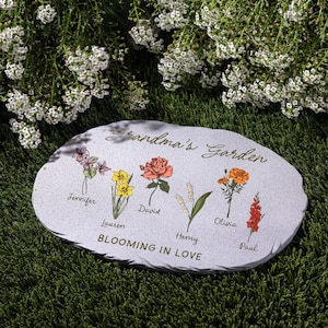 May include: A gray stone garden marker with the words "Grandma's Garden" and "Blooming in Love." It features colorful illustrations of flowers with names: Jennifer, Lauren, David, Henry, Olivia, and Paul. The stone is on green grass.
