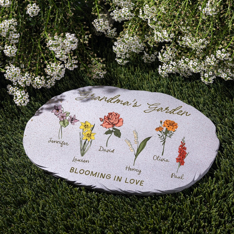 May include: A gray stone garden marker with the words "Grandma's Garden" and "Blooming in Love." It features colorful illustrations of flowers with names: Jennifer, Lauren, David, Henry, Olivia, and Paul. The stone is on green grass.