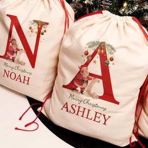 May include: Two personalized Christmas gift bags with red drawstring closures. The bags are made of natural canvas fabric and feature a red letter, a Santa Claus illustration, and the text "Merry Christmas" and a name. The bags are "N" for Noah and "A" for Ashley.