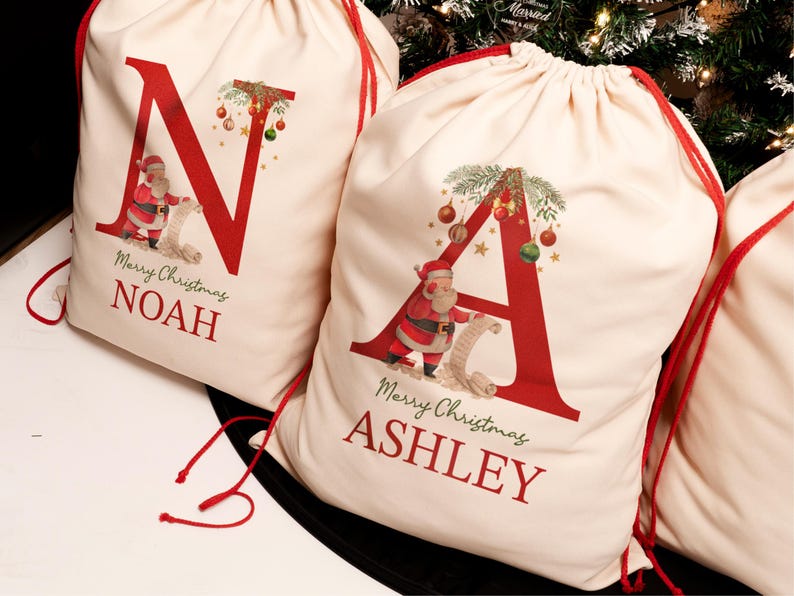 May include: Two personalized Christmas gift bags with red drawstring closures. The bags are made of natural canvas fabric and feature a red letter, a Santa Claus illustration, and the text "Merry Christmas" and a name. The bags are "N" for Noah and "A" for Ashley.