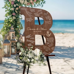 K&ouml;nnte beinhalten: Gro&szlig;es Hochzeitszeichen aus Holz in Form des Buchstabens "B" mit eingravierten Nachrichten, einem Anker und den Namen "Daria & Chris" mit dem Datum 09.12.2025. Das Schild steht auf einer Holzstaffelei, dekoriert mit Blumen, am Strand.