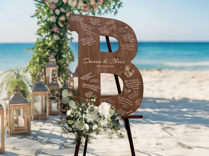 K&ouml;nnte beinhalten: Gro&szlig;es Hochzeitszeichen aus Holz in Form des Buchstabens "B" mit eingravierten Nachrichten, einem Anker und den Namen "Daria & Chris" mit dem Datum 09.12.2025. Das Schild steht auf einer Holzstaffelei, dekoriert mit Blumen, am Strand.