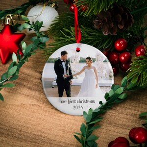 May include: Round white ceramic ornament with a photo of a couple in wedding attire standing in front of a floral arch. The text "our first christmas 2024" is printed below the photo.