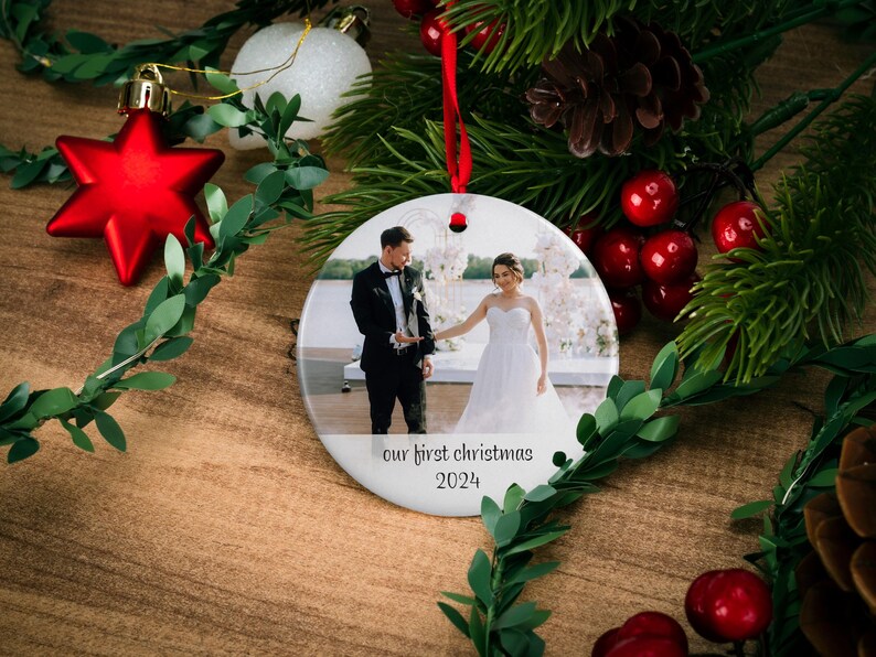 May include: Round white ceramic ornament with a photo of a couple in wedding attire standing in front of a floral arch. The text "our first christmas 2024" is printed below the photo.