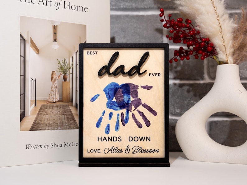 May include: A wooden plaque with a black frame and stand. The plaque reads "BEST dad EVER" in black raised letters. Below are blue and purple handprints and the words "HANDS DOWN LOVE, Atlas & Blossom."