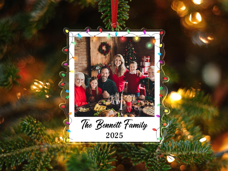 Pode incluir: Enfeite de Natal em acr&iacute;lico transparente com uma foto de fam&iacute;lia no interior, emoldurado por uma borda branca e uma sequ&ecirc;ncia de luzes coloridas. O texto "The Bennett Family 2025" est&aacute; impresso na parte inferior. O enfeite pende de uma fita vermelha.