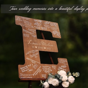K&ouml;nnte beinhalten: Holzschild in Form des Buchstabens "E" f&uuml;r eine Hochzeit mit handschriftlichen Nachrichten. Das Schild enth&auml;lt die Namen "Louis & Melisa" und das Datum "18. JUNI 2022". Wei&szlig;e Blumen und Gr&uuml;nzeug befinden sich am Fu&szlig; des Schildes. Der Text oben lautet: "Verwandeln Sie Hochzeitserinnerungen in ein sch&ouml;nes Ausstellungsst&uuml;ck."