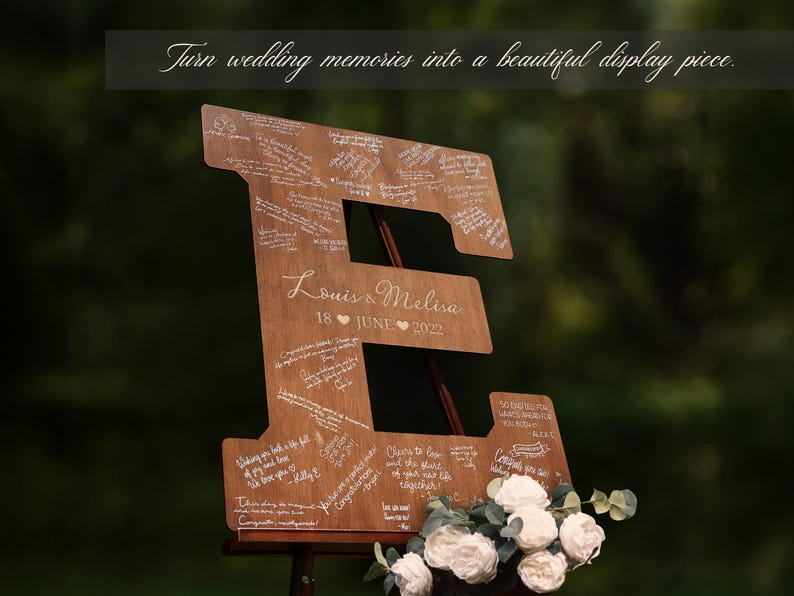 K&ouml;nnte beinhalten: Holzschild in Form des Buchstabens "E" f&uuml;r eine Hochzeit mit handschriftlichen Nachrichten. Das Schild enth&auml;lt die Namen "Louis & Melisa" und das Datum "18. JUNI 2022". Wei&szlig;e Blumen und Gr&uuml;nzeug befinden sich am Fu&szlig; des Schildes. Der Text oben lautet: "Verwandeln Sie Hochzeitserinnerungen in ein sch&ouml;nes Ausstellungsst&uuml;ck."